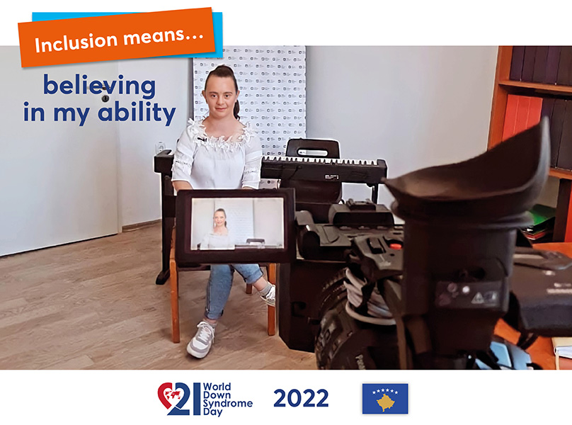 A woman with Down syndrome is sitting in a small studio for an interview. Around her are technical devices, including a monitor. Behind her is an electric piano and a shelf can be seen at the edge of the picture.