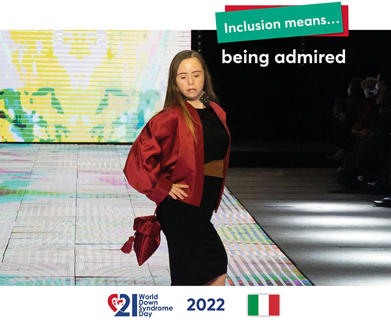 A young woman with Down syndrome stands confidently on a stage in a chic outfit with a shoulder bag.